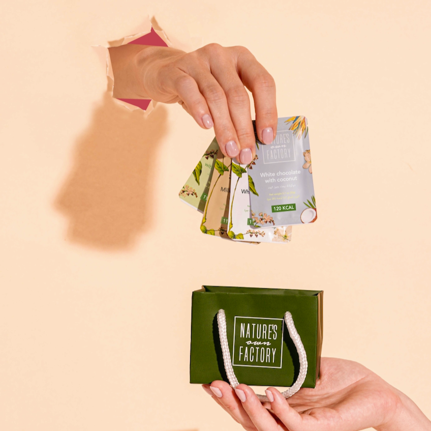 Hand placing mini products into a green 'Nature's own Factory' mini chocolate set bag on a beige background