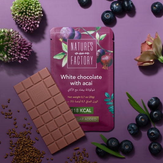 Nature’s Own Factory White Chocolate with Acai bar and packaging on a purple background, surrounded by blueberries and flowers. no added sugar, 118 kcal healthy chocolate.
